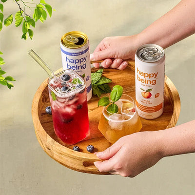 Hands holding a wooden tray with two Happy Being cans, a glass of berry-infused drink with blueberries, and a small dish of mint leaves, outdoors near greenery