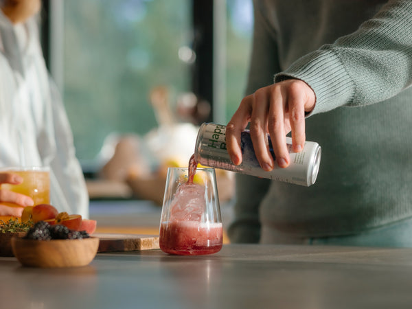 Glass of iced blueberry white tea being poured from a can, surrounded by fresh berries and fruit on a table, creating a refreshing wellness-focused setting.