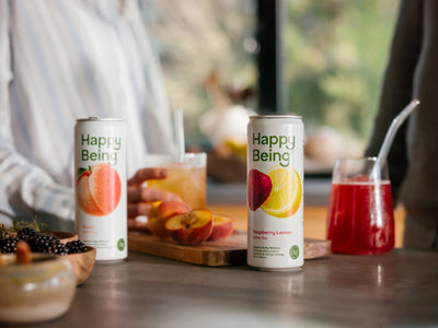 Happy Being Peach and Raspberry Lemon white tea cans with fresh peaches and a glass of Raspberry Lemon tea on a wooden tray.