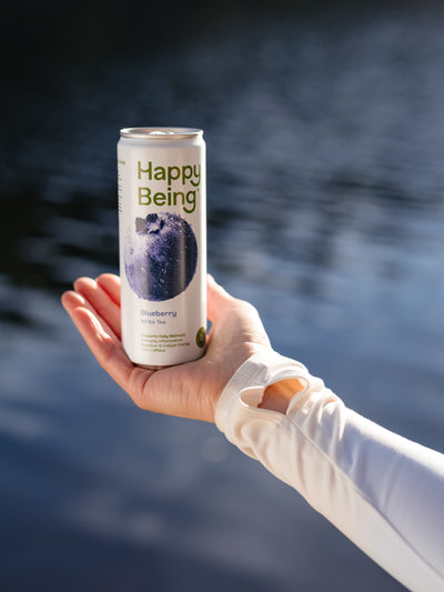 Hand holding a can of Happy Being Blueberry Sparkling White Tea against a calm water background