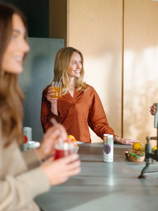 Woman smiling while holding a glass of Happy Being white tea drink with branded cans in the background