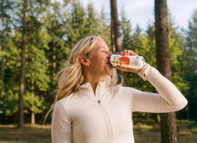 Woman enjoying a can of Happy Being Peach White Tea while standing outdoors in a sunny forest.