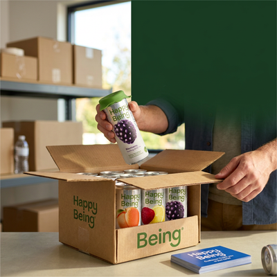 Hands unpacking a Happy Being can from an open shipping box filled with assorted flavor cans, warehouse boxes in the background
