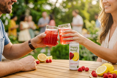 Two people toasting with glasses of pink Happy Being wellness beverage at an outdoor gathering, with a Happy Being can and fresh raspberries and lemons on the table