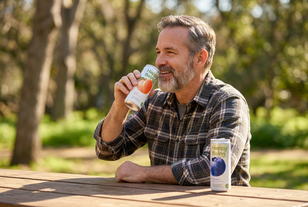 Man drinking Happy Being beverage outdoors at a wooden table, smiling in sunlight