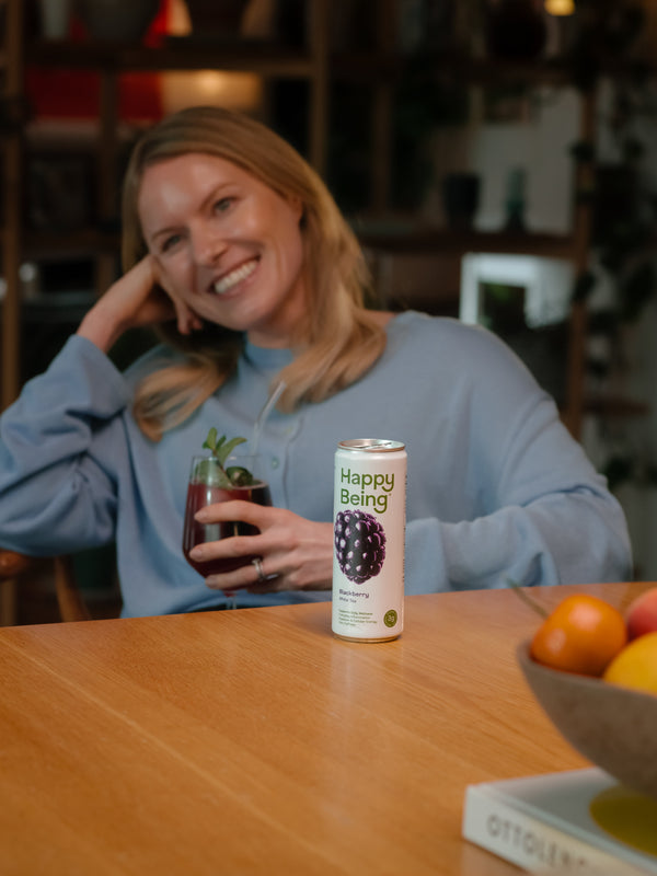 Woman holding a glass of Happy Being blackberry white tea beside an opened can of the drink outdoors.