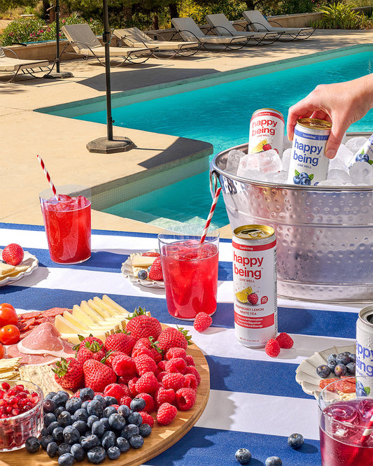 A picnic mat by the pool featuring Happy Being White Tea cans with charcuterie, fruits, and wellness-focused snacks.
