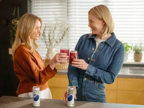 Two women smiling and toasting with glasses of Happy Being, with cans of Blueberry and Raspberry Lemon in the foreground.