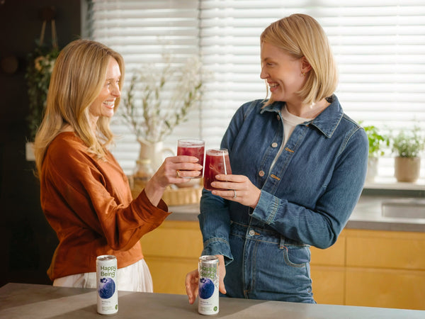 Two women smiling and raising glasses of Happy Being while Blueberry and Raspberry Lemon white tea cans sit in the foreground.