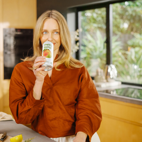 Woman smiling while drinking a can of Happy Being in a bright kitchen setting.