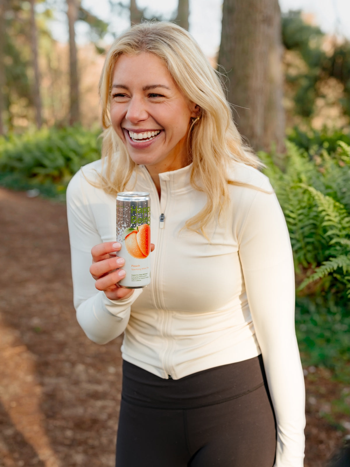 A smiling blonde woman outdoors in a wooded setting, wearing a fitted cream long-sleeve athletic top and dark leggings while holding a beverage can.